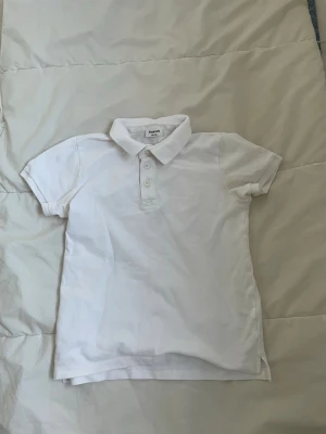 Vit pikétopp från Kappahl - Säljer en klassisk vit pikétopp från Kappahl i storlek 146/152. Toppen har korta ärmar, krage och tre knappar framtill. Materialet är mjuk bomull och modellen är normal passning. Perfekt till sommaren och enkel att matcha med jeans eller shorts.