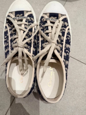 Christian Dior blå sneakers med mönster - Säljer ett par snygga Christian Dior sneakers i marinblått och beige med tydligt Dior-mönster. Skorna har rund tå, vit platt sula och snörning med logotyp på skosnörena. Materialet är textil och detaljerna är riktigt lyxiga. Perfekt för dig som gillar designer och streetstyle.