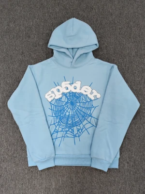 Blå hoodie från Sp5der  - Säljer en ljusblå hoodie från Sp5der med stor huva och ribbade muddar. Framsidan har ett coolt spindelnätsmönster i blått och ett stort vitt Sp5der-tryck med glittriga paljetter runt bokstäverna. Perfekt för dig som gillar streetwear och unika detaljer. Kom med bud gärna 