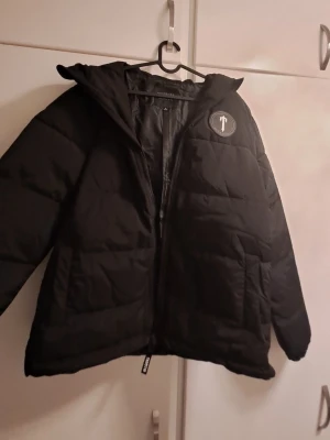 Svart pufferjacka från Trapstar - Säljer en svart pufferjacka med huva och dragkedja. Jackan har ett stort "Trapstar" tryck på ryggen och en rund patch på bröstet. Fylld för extra värme och har en clean, oversized look. Perfekt för kalla dagar och streetwear-stil.