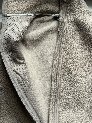 Beige teddyjacka med dragkedja - Mysig beige teddyjacka från lager med hel dragkedja framtill och två sidofickor med dragkedja. Jackan har en hög krage och är tillverkad i mjukt fleece-material som håller dig varm. Perfekt för kyliga dagar och har en clean, enkel look.