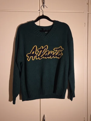 Mörkgrön stickad tröja Divided S - Säljer en mörkgrön stickad tröja från Divided i storlek S. Tröjan har ett stort gult och svart Atlanta-tryck på bröstet, v-ringad hals och ribbade muddar vid ärmslut och nederkant. Materialet är 100% akryl och modellen är långärmad med normal passform.