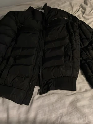 Tommy Hilfiger vinterjacka - Säljer en svart pufferjacka från Tommy Hilfiger med ribbade muddar och två bröstfickor med dragkedja. Jackan har en snygg quiltad design och klassisk logga på ena fickan. Perfekt för kalla dagar och har en cool, modern look. Helt oanvänd!