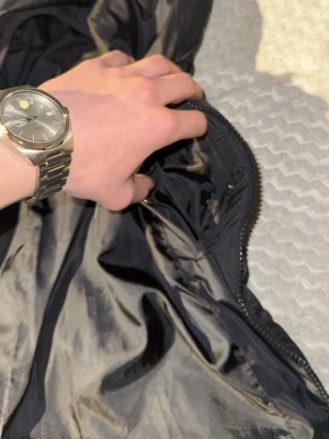 Svart dunväst från Lyle & Scott Junior - Säljer en svart dunväst från Lyle & Scott Junior med gul broderad logga på bröstet. Västen har quiltad design, dragkedja framtill och är tillverkad i syntetmaterial. Perfekt för dig som vill ha en stilren och varm väst med snygga detaljer.