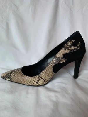 Custommade pumps med ormskinnsmönster - Säljer ett par snygga pumps från Custommade med spetsig tå och hög klack på 9 cm. Skorna har ett beige och svart ormskinnsmönster i flames-mönster på sidan och bakre del i svart mocka. Använd någon enstaka gång så i mycket gott skick, men såklart lite repor under sulan. 