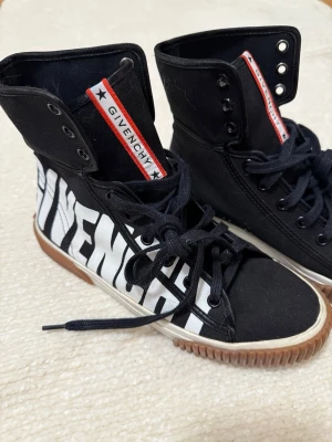 Svarta high-top sneakers från Givenchy - Säljer ett par svarta high-top sneakers från Givenchy med vit logotyp på sidan och röd detalj på plösen. Skorna har snörning, gummisula i brunt och insida i skinn. Snygg kontrast mellan svart och vitt, perfekt för dig som gillar statement pieces.