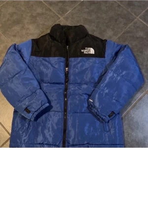 Blå och svart pufferjacka från The North Face - Säljer en blå och svart pufferjacka från The North Face med hög krage och tydlig logga på bröstet. Jackan har lång ärm, dragkedja framtill och är tillverkad i syntetmaterial. Perfekt för kalla dagar och har en schysst oversized vibe.