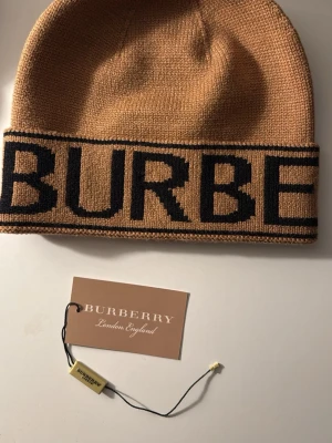 Burberry beige stickad mössa - Snygg beige stickad mössa från Burberry med bred svart logotyp runt om. Mössan har en klassisk form och är tillverkad i mjukt material som känns skönt mot huvudet. Perfekt för dig som vill ha en stilren accessoar med tydlig branding.