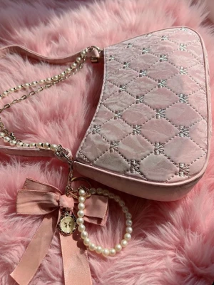 Rosa quiltad axelväska med pärldetaljer - Superfin rosa axelväska i sammet med quiltat mönster och silverdetaljer. Väskan har en kedja med pärlor och en stor rosett i satin samt en söt berlock. Perfekt för dig som gillar gulliga accessoarer och vill sticka ut med en unik look.