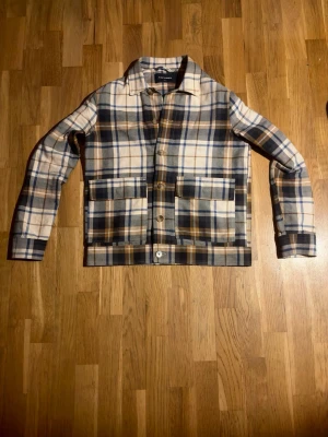 Rutig overshirt från A Day's March - Snygg rutig overshirt från A Day's March i beige, blå och bruna toner. Jackan har klassisk krage, stora fickor framtill och stängs med knappar. Tillverkad i mjuk bomull, perfekt för lager-på-lager och chill stil.