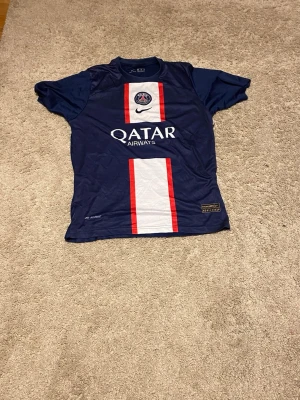 PSG Mbappé Nike matchtröja - Säljer en officiell Paris Saint-Germain matchtröja från Nike med Mbappé och nummer 7 på ryggen. Tröjan är marinblå med vita och röda ränder framtill, PSG-logga och Qatar Airways tryck. Perfekt för dig som älskar fotboll och vill ha en snygg supportertröja.