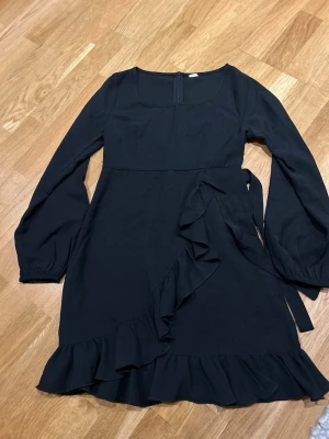 Svart omlottklänning från Shein Teen - Supersnygg svart omlottklänning från Shein Teen med långa ärmar och volangdetaljer längs nederkanten och omlotten. Klänningen har en markerad midja och knytband i sidan för extra stilpoäng. Perfekt för dig som gillar trendiga och feminina plagg.