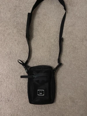 Svart axelväska från Acne Studios - Säljer en svart axelväska från Acne Studios med justerbar rem och praktisk dragkedja framtill. Väskan har ett meshfack på baksidan och ett ikoniskt ansiktsmärke på framsidan. Materialet är slitstark syntet och designen är stilren och modern.