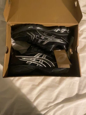 Svarta Asics sneakers - Säljer ett par svarta Asics sneakers med silvriga ränder på sidorna och mesh-överdel. Skorna har snörning och en platt sula som ger bra komfort. Perfekta för dig som gillar sportig stil och vill ha något som funkar till träning eller chill.