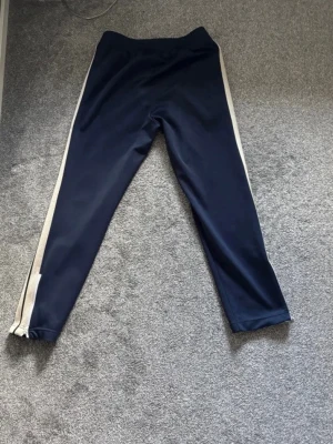 Palm Angels blå träningsbyxor - Säljer ett par mörkblå träningsbyxor från Palm Angels med vita sidoränder och logga på vänster ben. Byxorna har resår i midjan och raka ben. Materialet är mjukt och stretchigt, perfekt för en sportig look.