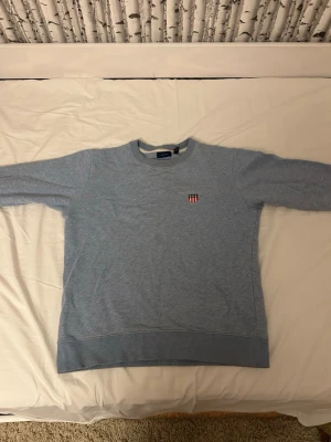 Ljusblå sweatshirt från GANT - Säljer en ljusblå sweatshirt från GANT med broderad logga och emblem på bröstet. Tröjan har rund hals, ribbade muddar vid ärmslut och nederkant samt är långärmad. Materialet är mjuk bomull, perfekt för chill dagar.