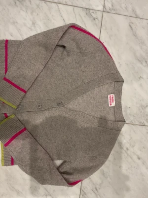 Beige kofta från From Future - Säljer en beige stickad kofta från From Future med knappar framtill. Koftan har ribbade muddar med detaljer i neonrosa och limegrönt, vilket ger en cool kontrast. Perfekt för dig som gillar stilrena plagg med färgglada inslag.