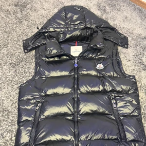 Svart dunväst från Moncler med huva - Snygg svart dunväst från Moncler med glansig finish, avtagbar huva och två dragkedjefickor framtill. Västen har klassisk Moncler-logga på bröstet och quiltad design. Perfekt för lager-på-lager och streetstyle.