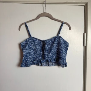Blå croppad linnetopp med volang - Söt blå croppad linnetopp med vitt småmönster, smala axelband och volangkant nertill. Toppen har knytning framtill och är perfekt för varma dagar. Lätt och luftig känsla, passar till både jeans och kjol. Oanvänd.