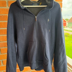 Ralph lauren zip hoodie - Mörkblå hoodie från Polo Ralph Lauren med dragkedja, huva med snörning och klassisk logga broderad på bröstet. Tröjan har känguruficka framtill och är tillverkad i mjuk bomullsblandning. Perfekt för chill dagar och snygg streetstil.                   Nypris: 2500kr.                                                                   Pris kan diskuteras