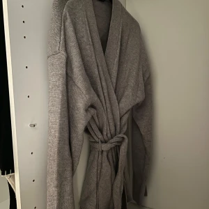 Grå stickad kofta med knytband - Mysig grå stickad kofta med omlottmodell och knytband i midjan. Koftan har långa ärmar och en avslappnad passform, perfekt att slänga på sig när det är kyligt. Enkel och stilren design som funkar till det mesta.