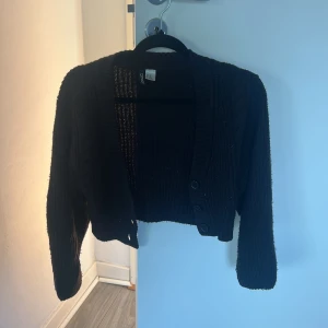 Svart croppad kofta från hm - Svart stickad kofta från hm med croppad passform och bred ribbad kant. Tröjan har långärmade ärmar, v-ringning och tre svarta knappar framtill. Perfekt att slänga över en t-shirt för en chill look.