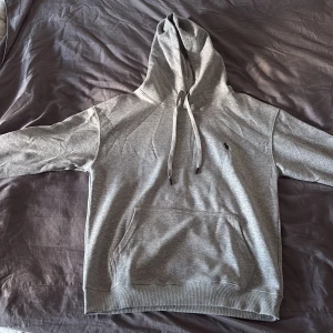 Grå hoodie från Polo Ralph Lauren - Snygg grå hoodie från Polo Ralph Lauren med klassisk svart logga på bröstet. Tröjan har huva med snörning, stor magficka och ribbade muddar vid ärmslut och nederkant. Mjuk bomullskvalitet som är perfekt för chill dagar.                                             Den är lite liten i storlek så jag skulle rekommendera Medium 
