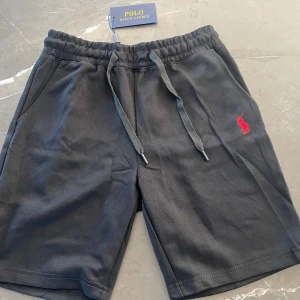 Svarta shorts från Polo Ralph Lauren - Svarta shorts från Polo Ralph Lauren med klassisk röd logobrodyr på vänster ben. Mjukt bomullsmaterial och bekväm midja med snörning. Perfekta för chill dagar och har två sidofickor. Snygg och enkel design som funkar till det mesta.