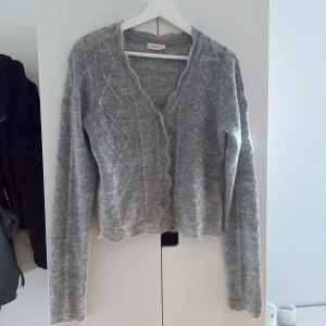 Grå stickad kofta från Nelly - Supermysig grå stickad kofta från Nelly med snygga knappar framtill. Koftan är croppad med lång ärm och har en vågig kant nertill för en trendig look. Perfekt att slänga över en topp när det är kyligt ute.