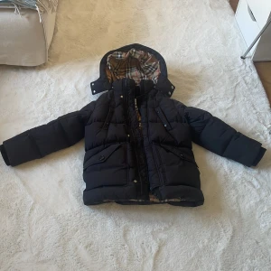 Svart dunjacka från Burberry med huva - Svart pufferjacka från Burberry med klassiskt rutigt foder i beige, rött och svart. Jackan har huva, dragkedja och knappar framtill samt flera fickor. Perfekt för kalla dagar och riktigt snygg med den ikoniska Burberry-looken. Stor i strl, står 128 /8 år men funkat på mig 10 år. 