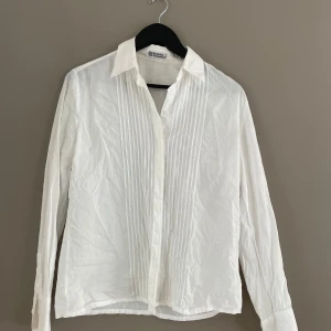 Vit skjorta från Nordiska Kompaniet - Klassisk vit skjorta från Nordiska Kompaniet med lång ärm, knappar framtill och snygga veckade detaljer på bröstet. Skjortan har en rak passform och är tillverkad i tunn bomull som känns lätt och fräsch. Perfekt till jeans eller kostymbyxor.