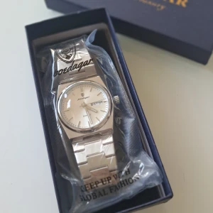 Silverklocka  - Stilren klocka från Poedagar med silverfärgat armband i metall och klassisk rund urtavla. Klockan har dag- och datumvisning samt quartz-urverk. Länkarna är breda och ger en modern känsla. Perfekt accessoar för dig som gillar en clean look.