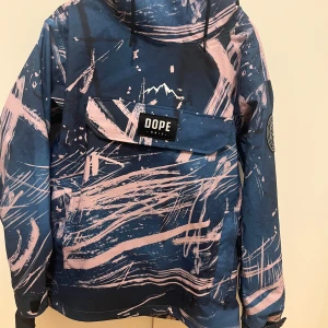 Blå och rosa vindjacka från Dope - Säljer en riktigt snygg vindjacka från Dope med blå bas och rosa abstrakt mönster. Jackan har stor magficka med Dope-logga, huva med dragsko och patch på ärmen. Ordinarispris är 2300kr.