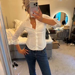 Vit figursydd skjorta med knappar - Säljer en klassisk vit skjorta med figurnära passform och knappar framtill. Skjortan har lång ärm och krage, perfekt för en clean och stilren look. Materialet känns mjukt och lätt, troligen bomull. Passar dig som gillar en enkel men snygg stil.