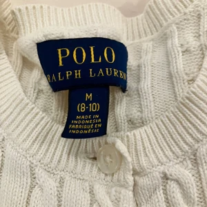 Vit kabelstickad kofta från Polo Ralph Lauren - Klassisk design med diskret rosa logga broderad på bröstet. Cardigan är i bra skick förutom ett litet defekt på höger arm (baksidan)..därav priset…men syns knappt och smutsen kan eventuellt tvättas bort. Storlek M 8-10, 140cm men känns ganska liten.