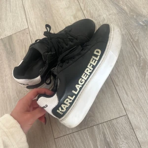 Karl Lagerfeld svarta sneakers - Svarta sneakers från Karl Lagerfeld med vit, grov sula och tydlig logga i guld på sidan. Skorna har snörning och vit häl med strukturmönster och svart logotyp. Tillverkade i skinnliknande material och har en chunky, modern look. Går säkert att få finare med en tvätt.