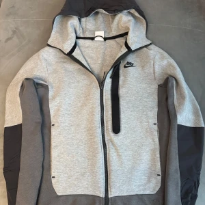 Grå Nike hoodie med dragkedja XS - Nike hoodie i ljusgrått och mörkgrått med svart logga på bröstet. Tröjan har hel dragkedja, huva och en svart bröstficka med dragkedja. Tillverkad i mjuk bomullsblandning, perfekt för chill eller träning. Liten deffekt längst ner inget som syns mycket :)