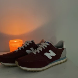 Vinröda New Balance COMP 100 sneakers - Snygga vinröda New Balance COMP 100 sneakers med klassisk retrostil. Skorna har vit N-logga på sidan, vita snören och detaljer i grå mocka på hälen. Yttersulan är beige med blå rand och ovandelen är i textil och mocka. Perfekta för dig som gillar sportig och stilren vibe.