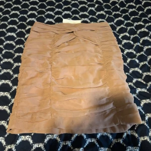 Beige brun kortkjol med volangdetaljer - Säljer en beige kortkjol med snygga volangdetaljer och omlottkänsla framtill. Kjolen har en markerad rosett i midjan och dragkedja bak.  Ny pris- 149kr.    Mitt pris 90kr