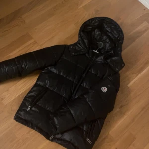 Svart Moncler pufferjacka med huva - Säljer en svart pufferjacka från Moncler med huva och dragkedja. Jackan har två fickor med dragkedja och klassisk Moncler-logga på bröstet. Materialet är glansigt och jackan är vadderad för extra värme. Perfekt för kalla dagar och riktigt snygg streetstil.