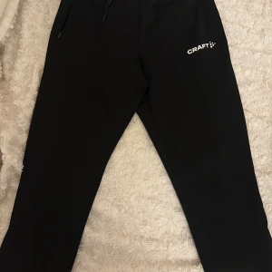 Svarta träningsbyxor från Craft XS - Svarta träningsbyxor från Craft i storlek XS. Byxorna har dragsko vid midja, dragkedjor vid bensluten och är tillverkade i ett mjukt och stretchigt material. Perfekta för träning eller chill. Snygg och enkel design med Craft-logga på låret.