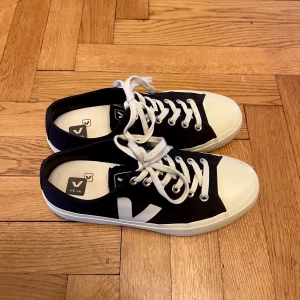 Svarta Veja sneakers - Snygga svarta Veja sneakers med vit tå och sula samt stora vita V-loggor på sidorna. Skorna har klassisk snörning och är tillverkade i canvas med gummidetaljer. Perfekta för dig som gillar stilrena och trendiga sneakers. Dom har bara använts några gånger. Tveka inte på att skriva om du har några frågor eller vill ha fler bilder!