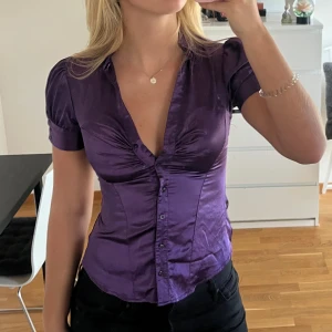 Lila glansig blus från River Island - Säljer en lila blus från River Island i storlek 34 med korta ärmar och knappar framtill. Blusen har v-ringning, figurnära passform och snygga rynkdetaljer vid bysten💗
