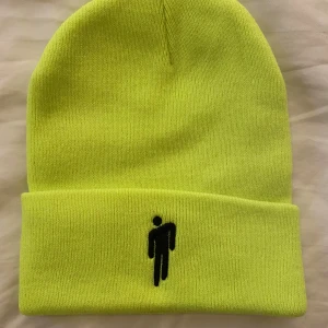 Billie Eilish neongrön mössa  - Neongrön mössa med uppvikt kant och svart broderad Billie Eilish-logga framtill. Tillverkad i mjukt, stickat material som håller dig varm och synlig. Perfekt för dig som vill sticka ut med en färgstark accessoar.