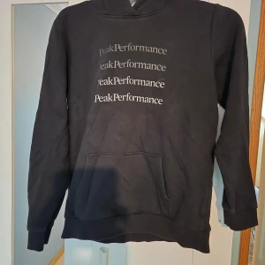 Svart hoodie från Peak Performance - Svart hoodie från Peak Performance med tryckt logga i flera rader på bröstet. Klassisk känguruficka framtill, ribbade muddar och huva. Perfekt för en avslappnad och sportig stil.