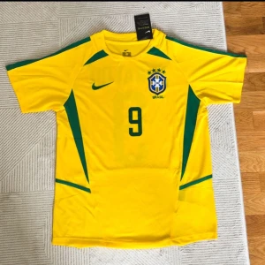 Brasilien Ronaldo 9 fotbollströja Nike - Gul Brasilien fotbollströja från Nike med gröna detaljer, nummer 9 och Ronaldo-tryck på ryggen. Klassisk design med korta ärmar, rund hals och broderat landslagsemblem på bröstet. Perfekt för dig som älskar fotboll och vill hylla en legend.
