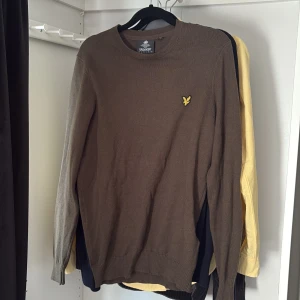 Brun stickad tröja från Lyle & Scott - Snygg brun stickad tröja från Lyle & Scott med klassisk rund hals och den ikoniska gula örnloggan på bröstet. Tröjan har långa ärmar och ribbade muddar för en clean och stilren look. Perfekt för lager-på-lager under hösten och vintern.