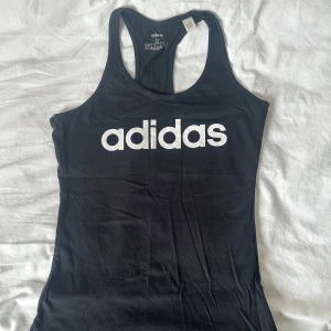 Svart träningsstopp från Adidas - Svart linnetopp från Adidas med stort vitt logotryck framtill. Modellen är ärmlös med brottarrygg och har en tight passform. Perfekt för träning eller en sportig look