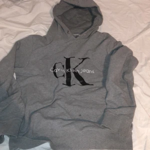 Grå hoodie från Calvin Klein  - Säljer en grå hoodie från Calvin Klein  med stor svart CK-logga och vit text på bröstet. Tröjan har huva och långa ärmar, perfekt för en chill och avslappnad stil.