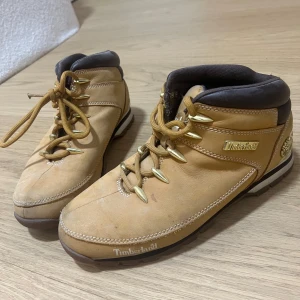 Beige Timberland kängor med snörning - Säljer ett par klassiska Timberland kängor i beige mocka med bruna detaljer och robust snörning med guldfärgade öljetter. Skorna har rund tå, platt sula och tydliga Timberland-loggor på sidan och hälen. Perfekta för dig som gillar street och outdoor.
